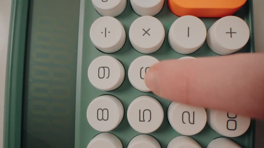 A person uses a green calculator to sum up values shown on a digital display next to a pen and highlighter on a white desk
