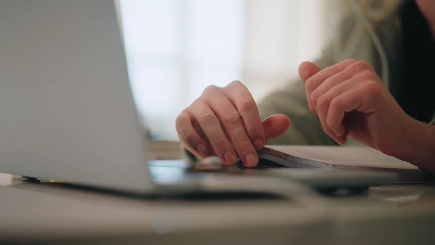 A womans hands rest on a white desk while she uses a laptop touchpad and keep her other hand near an open notebook in an indoor setting