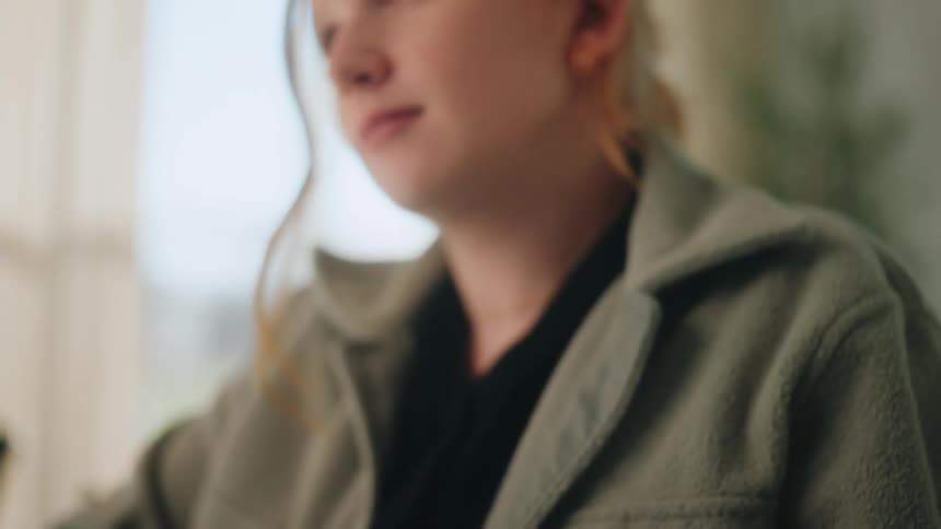 A redhaired woman sits at a desk with an open book and a pen looking up from her reading with a thoughtful expression as soft daylight illuminates her face in a quiet indoor setting
