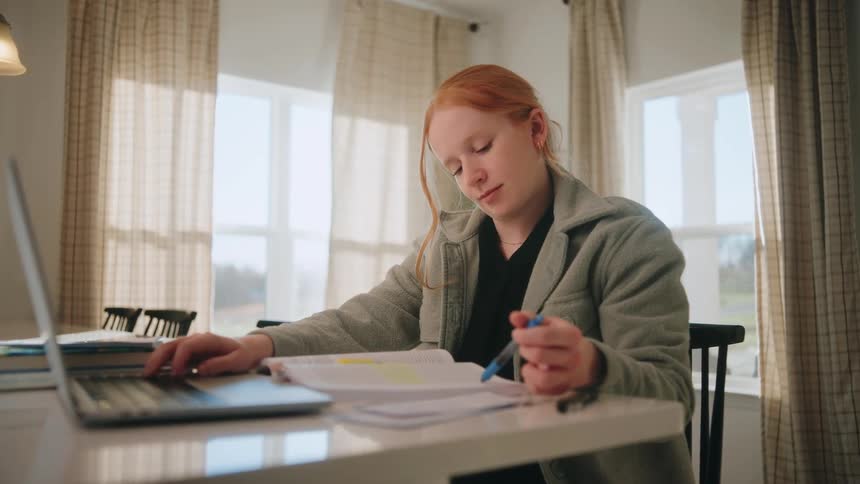 A focused teenager studies at a sunlit table with an open textbook and laptop in a bright room