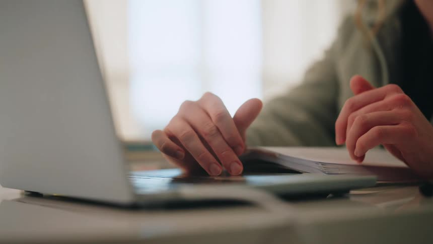 A woman sits at a white desk and begins typing on a laptop keyboard after resting her hands near a notebook