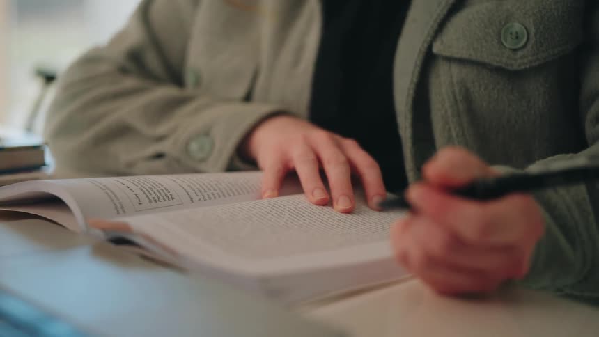 Young adult woman sitting while reading and writing on a book over a desk in front of a laptop computer indoors during day time