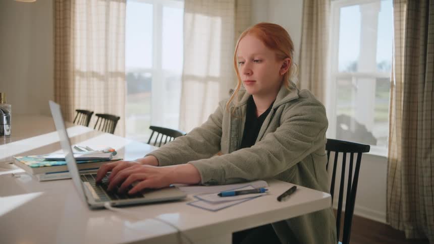 A young woman with red hair sits at a white table focused on typing on a silver laptop she wears a grey jacket and works in a bright room with natural light coming through large windows