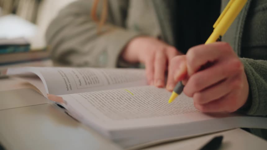 A woman wearing a grey jacket uses a yellow highlighter to mark specific lines of text in an open textbook resting on a white table