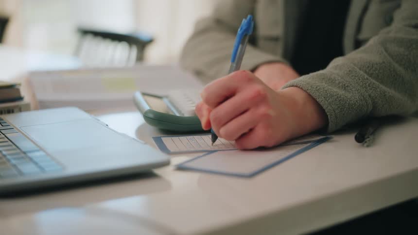 Person filling out a form at a desk with a pen calculator and laptop nearby