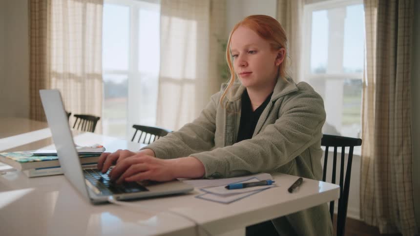 Redhaired woman typing on laptop in welllit room with books and notebooks on the table