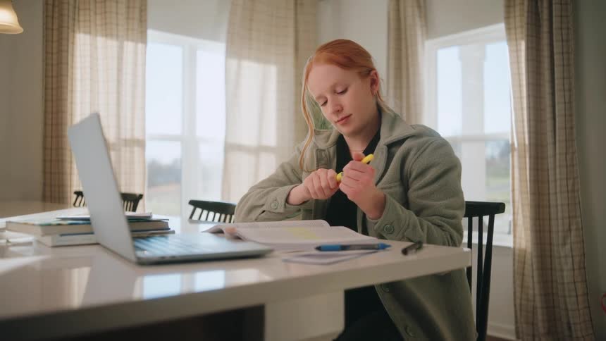 A young woman with red hair is sitting at a white table focused on her studies there is a laptop open to her left and several books stacked nearbyshe holds a yellow highlighter in her hands carefully removes the cap and begins to mark sections in a