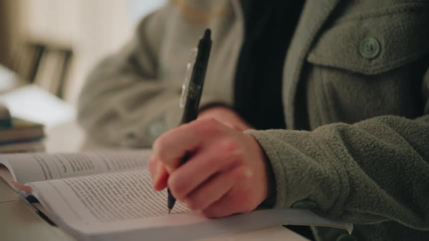 A woman with long red hair looks thoughtfully ahead before looking down to write in a notebook with a black pen while wearing a green fleece jacket in a bright indoor setting