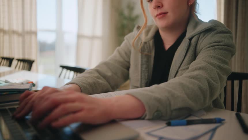 A teenage girl with red hair sits at a white dining table looking down at an open book before typing on her laptop while surrounded by school supplies and pens in a bright room