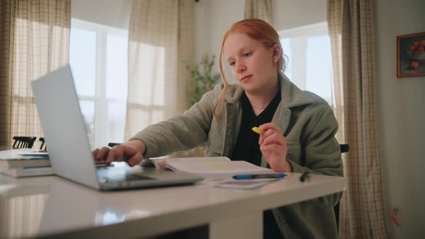 A focused young woman studies intently at a dining table in a sunlit room