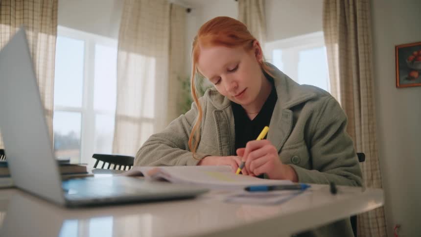 A girl highlighting on a textbook with a yellow pen while sitting in a house with a opened laptop on the table