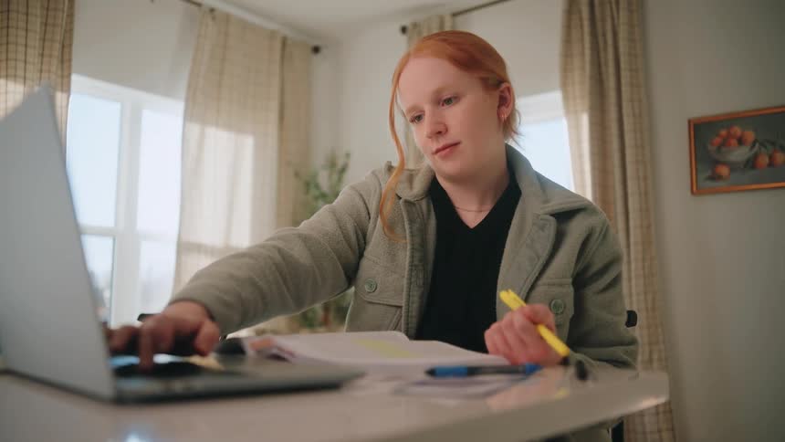 Person seated at a table uses a laptop while holding a highlighter and reviewing printed documents