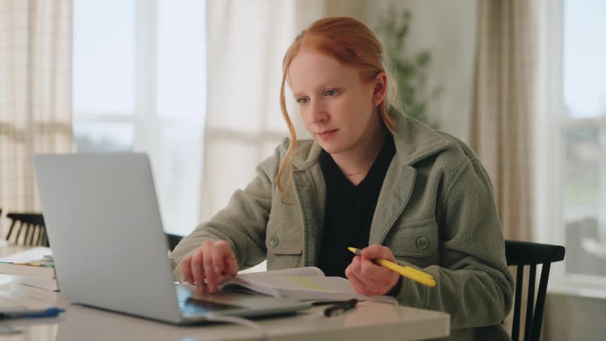 A young woman with red hair sits at a white table using a yellow highlighter on a textbook before looking at her laptop screen and scrolling with the trackpad and she wears a green fleece jacket in a bright sunlit room with curtains behind her