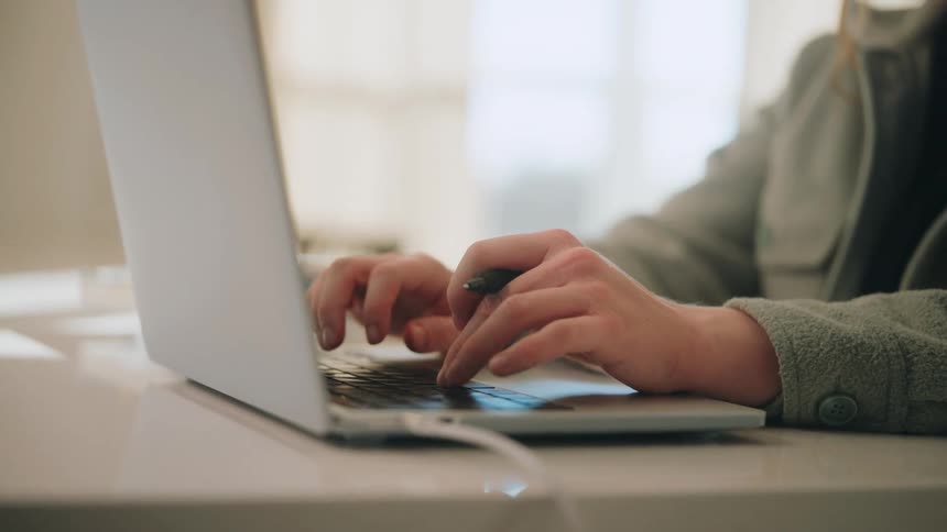 A woman wearing a gray jacket sits at a white table and types on a silver laptop while holding a black pen between her fingers
