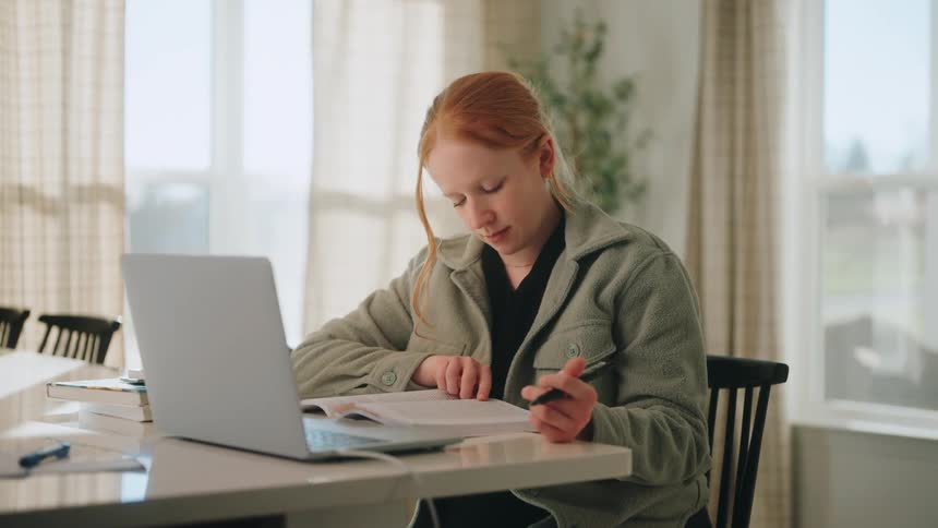 Person seated at a table reviews a notebook while using a laptop and holding a pen