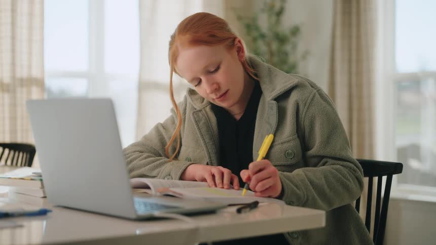 A teenage girl with red hair sits at a white desk in a bright room intently studying
