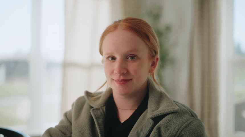 A young woman with red hair looks off to the side before turning to face the camera with a soft smile she is wearing a green fleece jacket and sits in a brightly lit room with a blurred window in the background