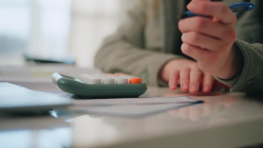 A person uses a green calculator to perform computations and then writes notes with a blue pen on a document while sitting at a desk with a laptop