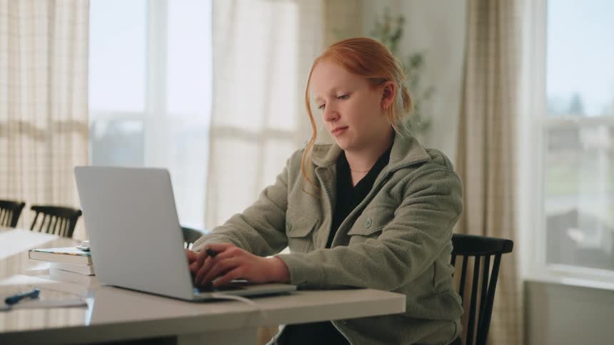 A focused student studying while typing on a laptop at a brightly lit table