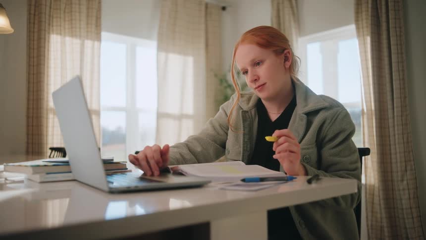 Person seated at a table uses a laptop while holding a highlighter with a notebook and pens resting on the surface