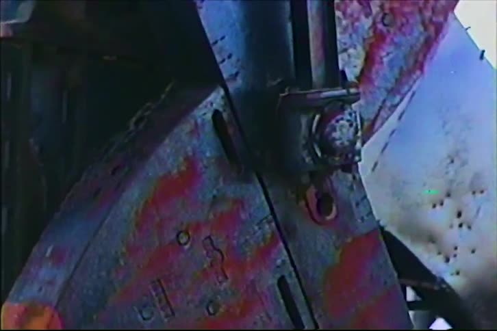 A closeup shot of large rusted industrial machinery moving slowly against a bright sky revealing weathered metal plates heavy bolts and mechanical components covered in red corrosion and aged blue paint