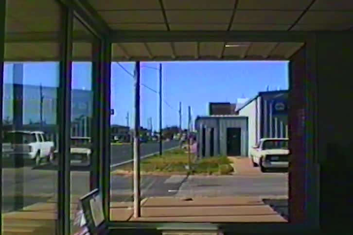 Urban sidewalk view seen through a storefront window with street utility poles parked vehicles and commercial buildings under a clear sky