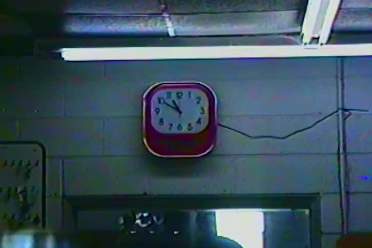 A red square analog clock hangs on a cinder block wall in a dimly lit workshop beneath a flickering fluorescent light fixture and a power cable