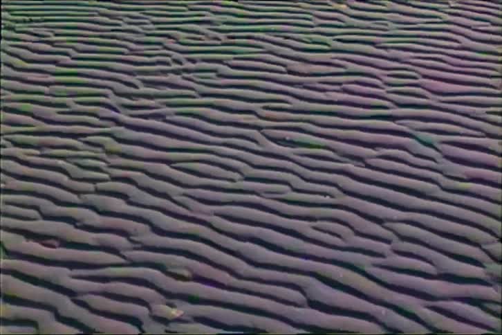The video shows a textured expanse of sand ripples covering the ground in a repetitive wavy pattern the surface is cast in deep shadows and highlights creating a rhythmic visual effect across the entire frame