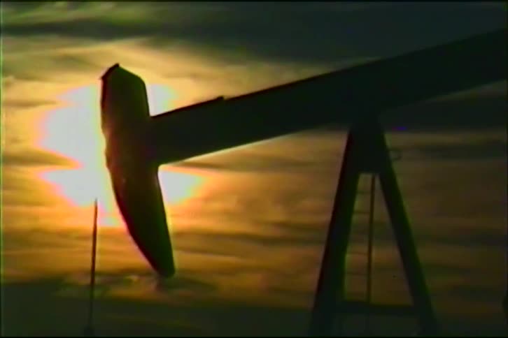 An oil pump jack operates rhythmically against the warm glow of a setting sun casting a dark silhouette across the horizonthe footage is captured in a vintage vhs style featuring heavy grain and scan lines that give the industrial landscape a nostalg