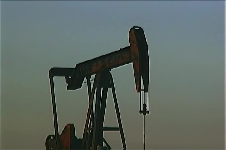 An industrial pumpjack operates in an open field during twilight its mechanical arm rhythmically lifting and lowering a polished rod to extract oil from the ground against a clear fading sky