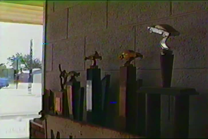 A row of carthemed trophies sits on a shelf against a cinder block wall inside a garage while an open doorway in the background reveals a bright outdoor parking area and distant buildings under a clear sky