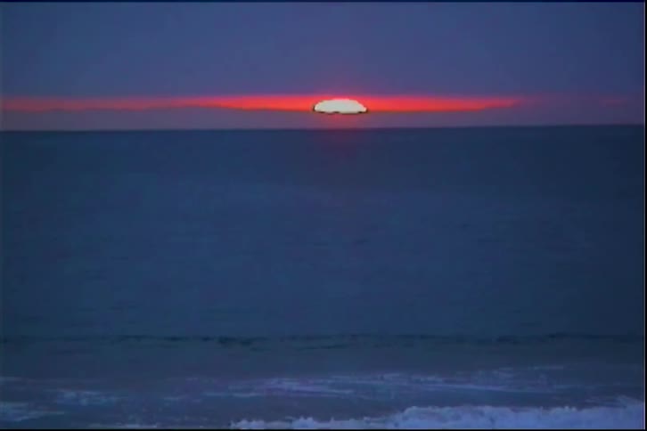 A vibrant orange sun partially dips below the dark horizon of a calm blue ocean with white waves crashing on a beach in the foreground under a deep blue sky at sunset