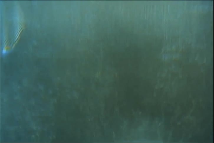 This video features a highly abstract textured sequence of light shimmering through a dark liquidlike environment the visual style mimics vintage film stock with heavy grain lens flares and a soft blueteal color palette