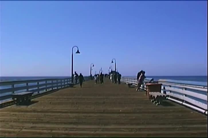 People are walking along a wide wooden pier that stretches out toward the ocean under a clear blue sky the pier is lined with benches and street lamps and several individuals are scattered along the walkway