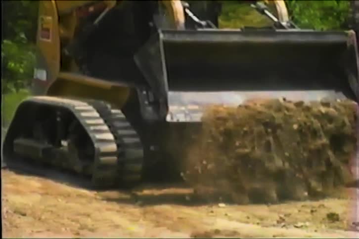 Tracked construction vehicle moves soil at outdoor site with greenery in background
