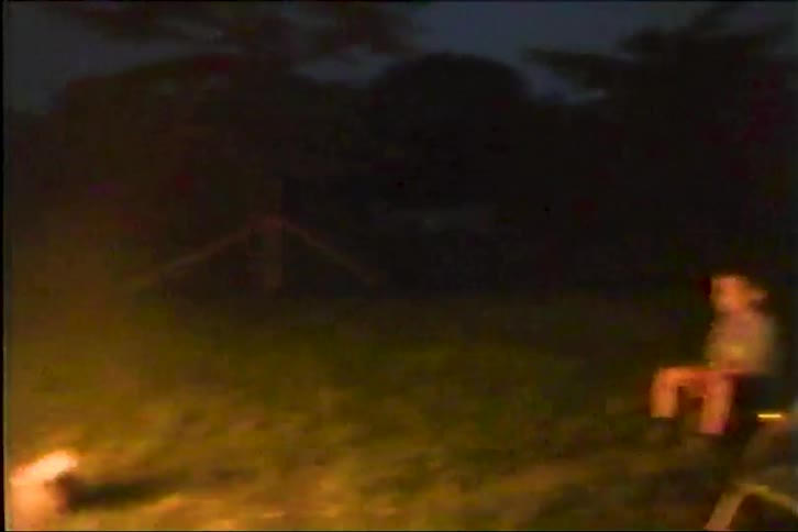 Three young boys sit on lawn chairs around a glowing campfire at night