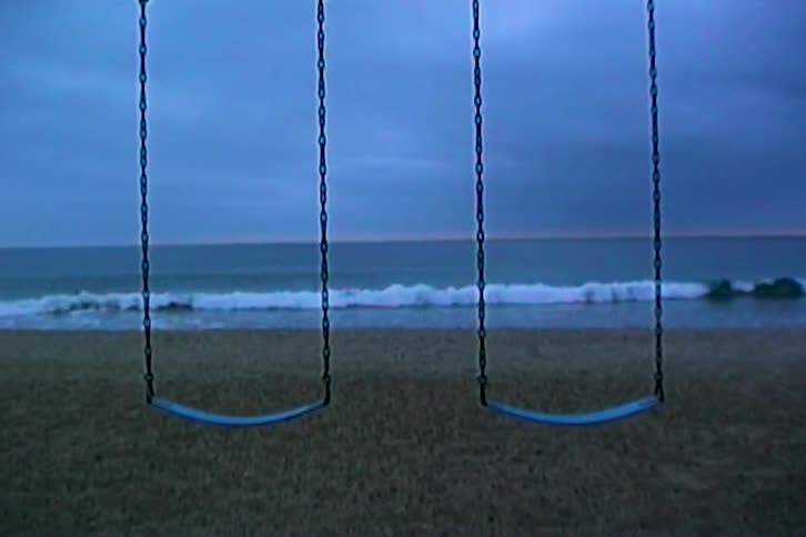 Two empty swings hang over beach sand with waves in the background under a cloudy blue sky