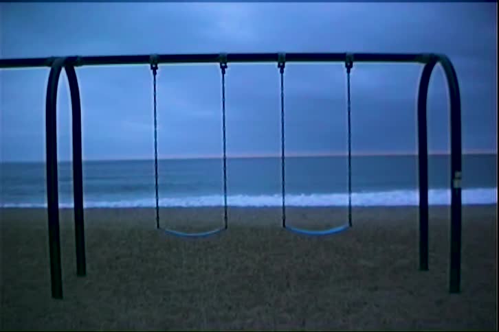 A stationary shot of a swing set on a beach during twilight as waves crash against the shore in the background against a overcast sky