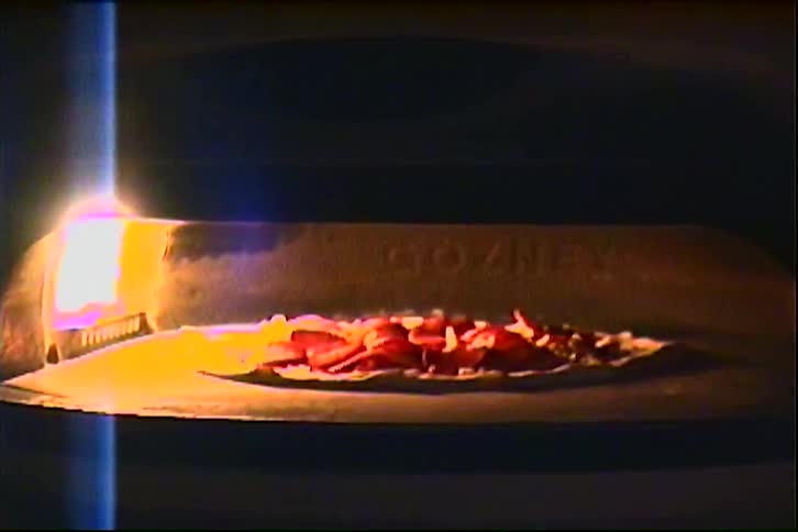 Closeup of a hand placing a pizza into a wood fired oven lit by open flames
