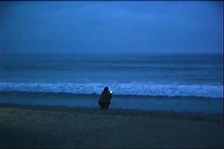 The video shows a person sitting alone on a beach during twilight facing the ocean as the waves roll in they hold up a phone to take a photo or video of the water