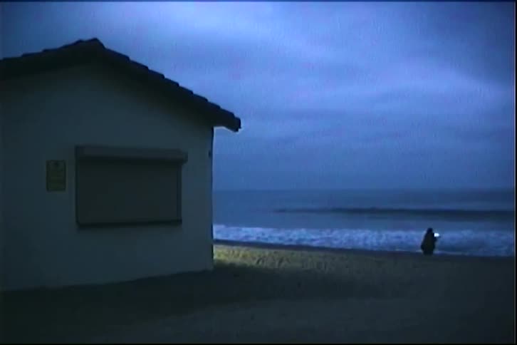 Person walks alone along the beach at dusk with the sea in the background and a small building on the left