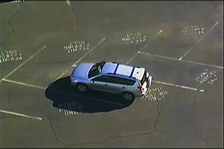 A silver suv is parked diagonally across multiple designated permit parking spaces in an asphalt lot the scene is captured from a highangle security camera viewpoint showing the vehicles shadow stretching across the cracked pavement
