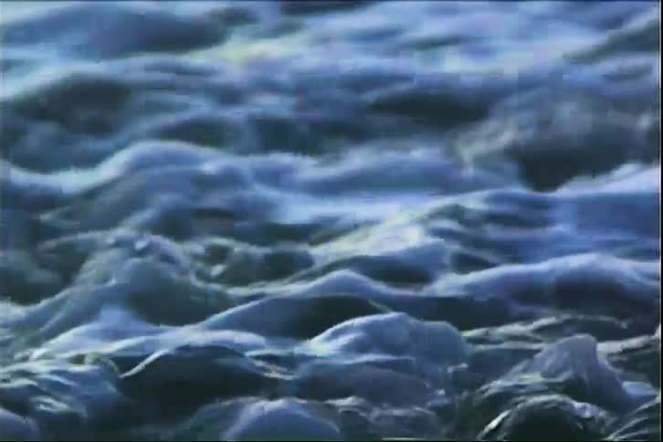 In a shaky closeup shot with a vintage film look turbulent blue waves and white foam crash over smooth shoreline rocks