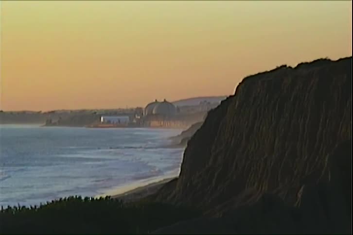 The video features a goldenhued coastline at dusk where gentle waves roll onto a sandy beach large dark cliffs stand in the foreground overlooking a distant shoreline dotted with industrial or coastal buildings