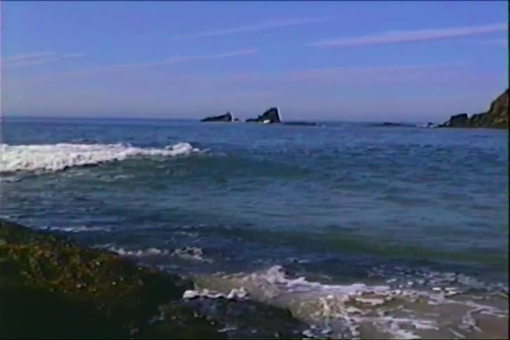 Waves breaking near a rocky shoreline with rock formations rising on the horizon under a blue sky