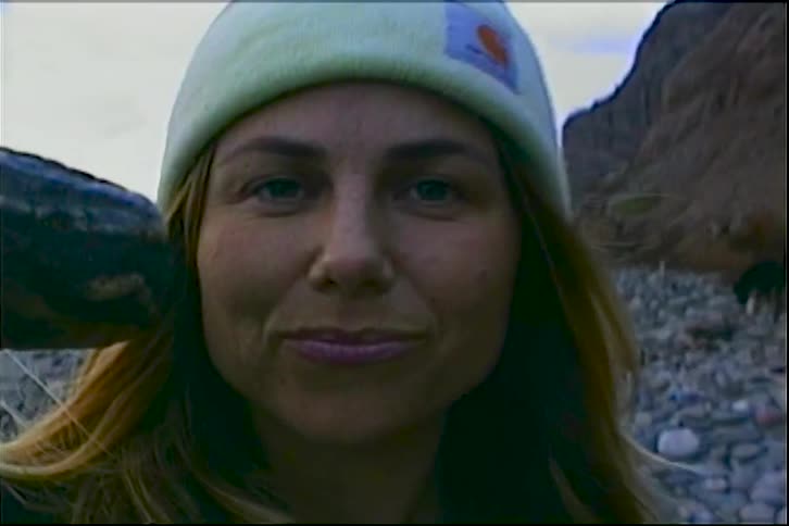 A woman in a beanie walks along a rocky beach and holds up a dark flat stone toward the camera the camera zooms in on her face as she smiles and looks directly into the lens while standing by the ocean