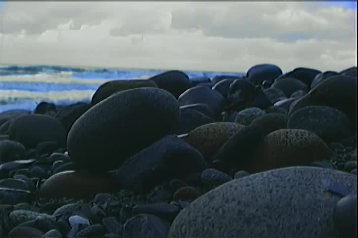 Smooth dark stones of various sizes cover the foreground of a coastal beachin the background light blue waves roll under a bright cloudy sky