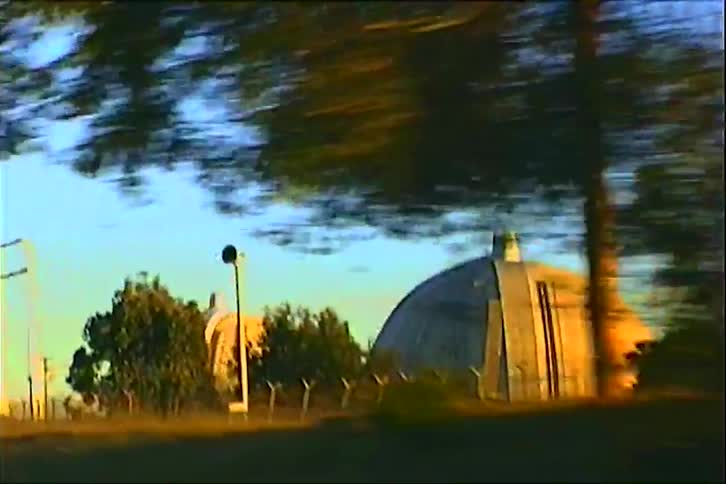 During a road trip at midday a building with an arched roof appears along the roadside while the road itself is lined with trees and power lines stretching into the bright distance