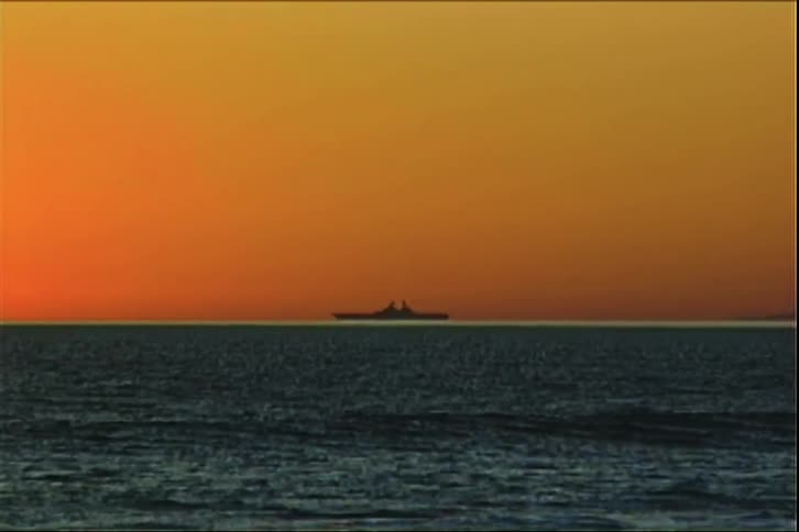 A large ship is silhouetted against a bright orange sky as it sits on the horizon of the ocean waves gently roll in the foreground under the warm glow of the setting sun