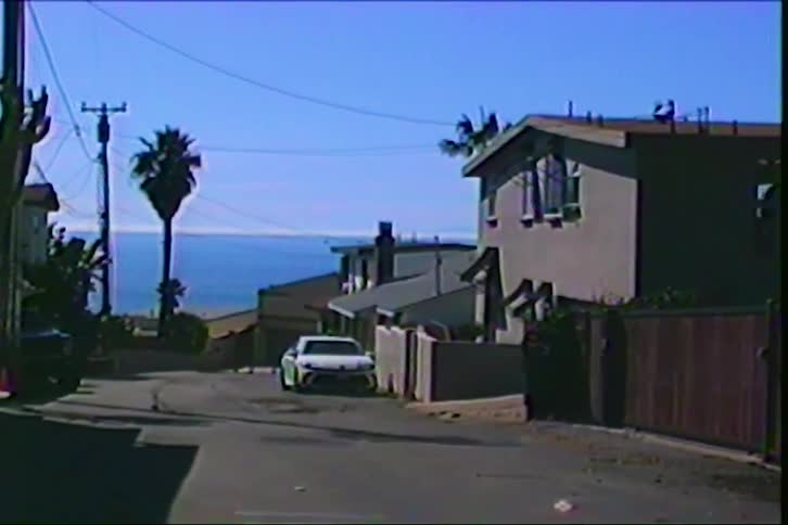 A street slopes down toward the blue ocean between residential houses under a clear sky featuring a white car parked on the roadside near a tall palm tree in a coastal neighborhood